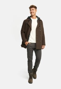 Cappotto doppiopetto marrone con cappuccio grigio, camicia color crema sotto, pantaloni grigi e stivali marroni con lacci. Design semplice, tessuto resistente.