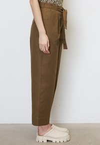 Pantalons marron à taille haute et coupe fuselée en coton, dotés d'une ceinture nouée et de plis sur les côtés. Associés à des chaussures slip-on beiges à semelle épaisse.