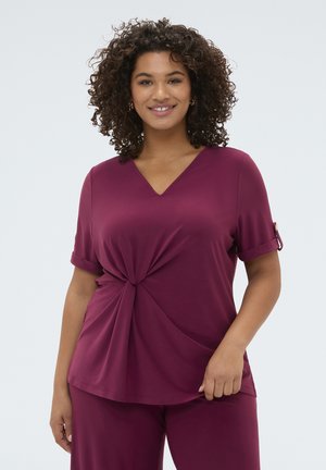 Lachende vrouw met krullend haar die een paarse top met korte mouwen en V-hals draagt, met een gedraaide voorkant en bijpassende broek.