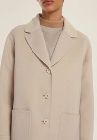 Cappotto beige, strutturato, con colletto a rever, tre bottoni e due tasche frontali. Realizzato in un tessuto morbido e testurizzato. Design elegante e minimale.