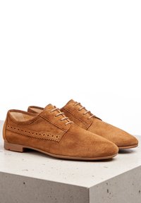 Chaussures oxford en daim marron avec une texture lisse, présentant des détails de brogue sur le côté et une semelle plate en caoutchouc.