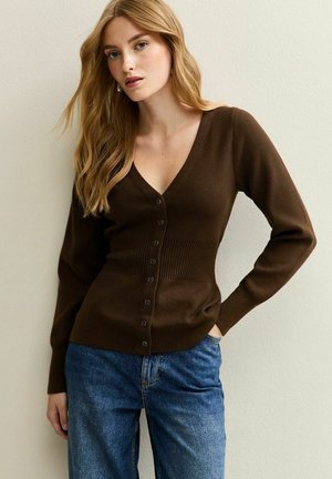 Cárdigan marrón con canalé, de escote en V y cierre de botones, con mangas largas y cintura ajustada. Lleva puesto sobre unos jeans de mezclilla azules.
