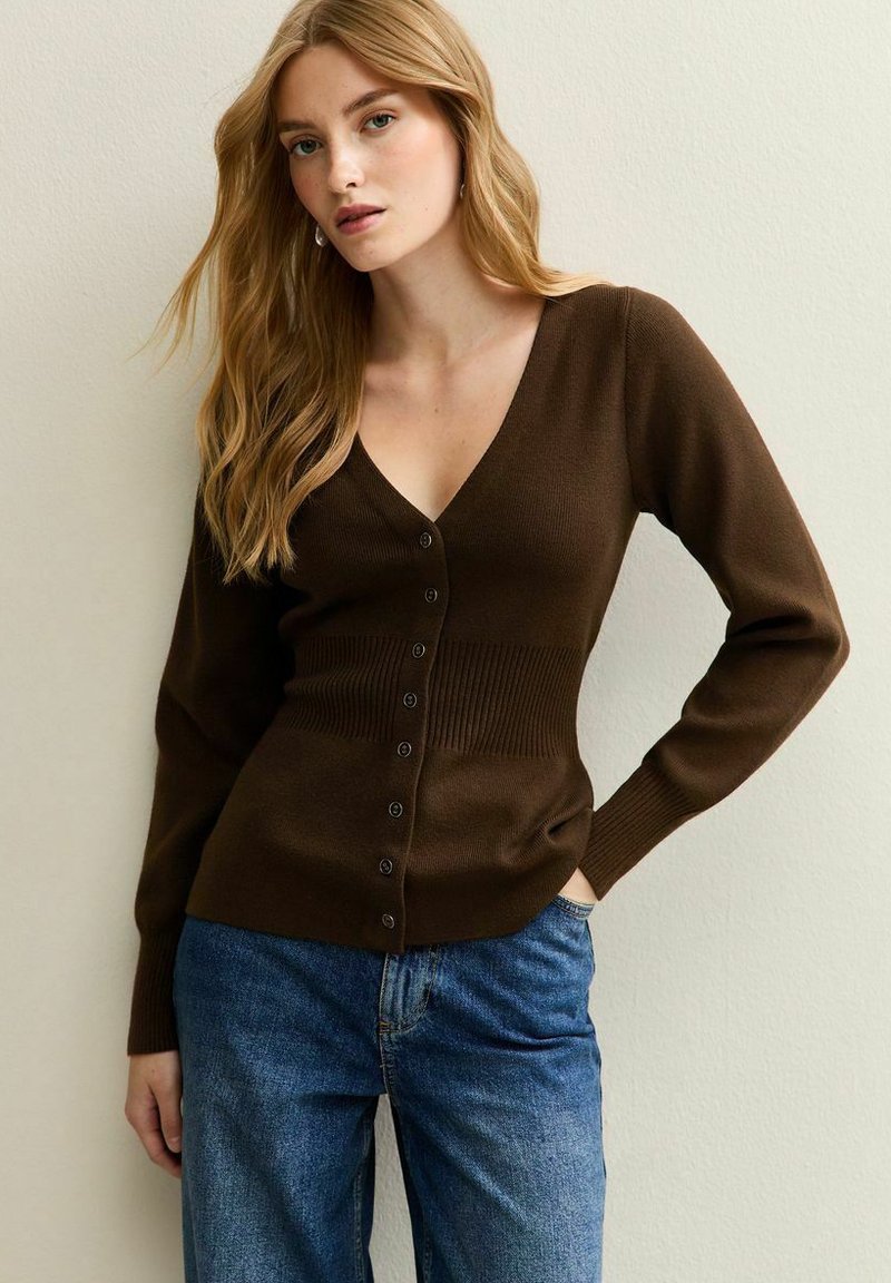 Cardigan marrone a coste con scollo a V e chiusura a bottoni, con maniche lunghe e vita aderente. Indossato sopra jeans in denim blu.