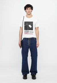Weißes T-Shirt mit schwarzem Grafikdruck und rotem Text, kombiniert mit lockeren blauen Jeans aus Denim mit Knopfdetails und einer schwarzen Umhängetasche.