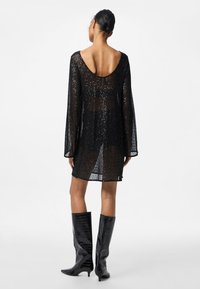 Vestido preto com lantejoulas, mangas compridas, costas baixas e textura transparente, combinado com botas pretas brilhantes até ao joelho.