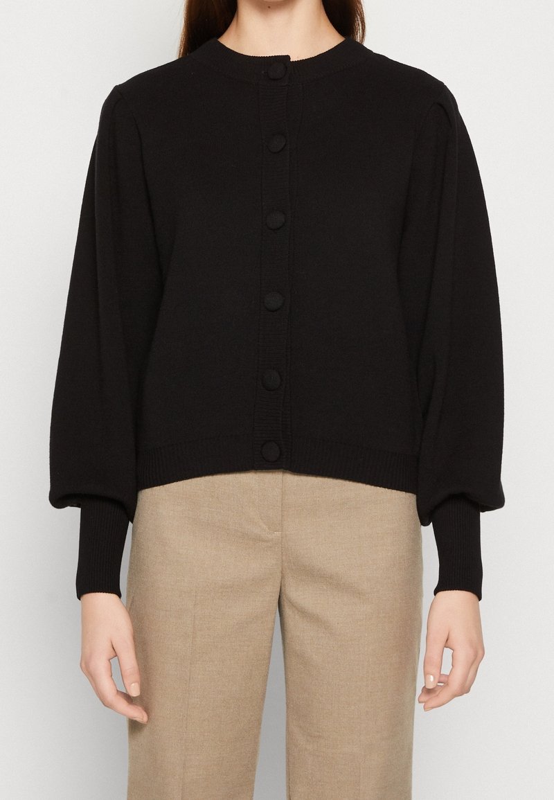 Femme portant un cardigan noir boutonné avec des manches bouffantes et un pantalon beige taille haute, debout devant un fond uni.