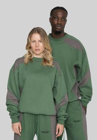 Zwei Models tragen passende grüne und graue Sweatshirts sowie Jogginghosen mit "Multiply Apparel" Branding vor einem einfarbigen grauen Hintergrund.