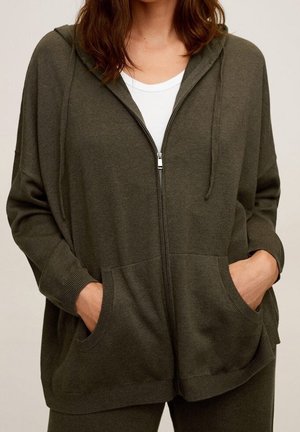 Kobieta ubrana w ciemnozieloną bluzę rozpinaną na zamek z przednimi kieszeniami, pod którą ma białą koszulę, ręce schowane w kieszeniach, stoi na jednolitym tle.