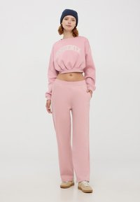 Felpa corta rosa con la scritta "PHOENIX", abbinata a pantaloni della tuta larghi e coordinati. Outfit realizzato in un tessuto morbido e testurizzato.