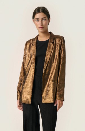 Sacou metalizat bronz, cu o textură netedă, cu rever notat și două buzunare frontale, purtat peste un top negru cu dungi și pantaloni plisati negri.