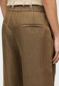 Καφέ λινό παντελόνι με χαλαρή εφαρμογή, που διαθέτει μια μοναδική πίσω τσέπη και διακριτική υφή, εμφανιζόμενο από γωνία με ένα χέρι να ακουμπά κοντά.