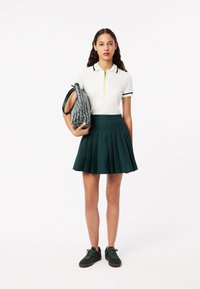 Polo blanc avec fermeture éclair et rayures vertes, jupe plissée vert foncé, et un sac à main gris à motifs. La mannequin porte des baskets vertes.