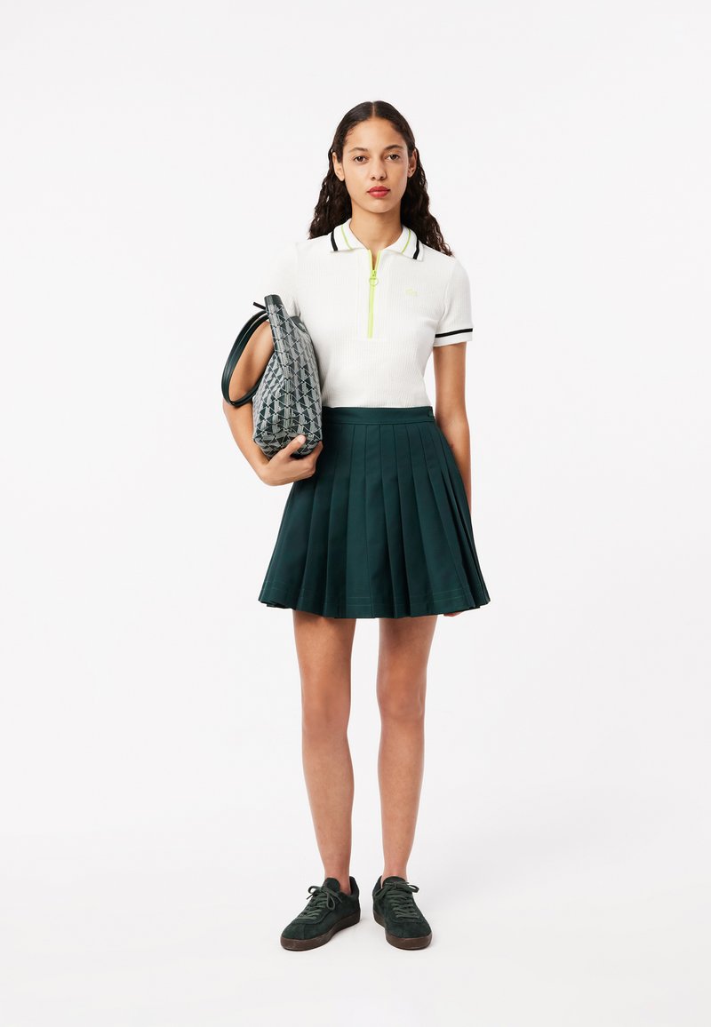 Weißes Polohemd mit grünem Reißverschluss und Streifen, dunkelgrünen Faltenrock und einer grauen gemusterten Handtasche. Das Model trägt grüne Sneakers.