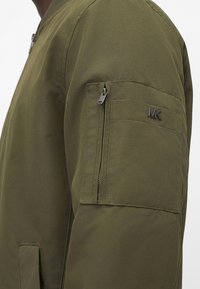 Κοντινό πλάνο στο μανίκι ενός ελιάς πράσινου μπουφάν με τσεπάκι με φερμουάρ και μεταλλικό σήμα "MK" στο πάνω μέρος του βραχίονα.