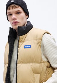 Beež pufferivest musta kraega, millel on sinine "HUGO" logo, tõmblukuga suletav ja selle all on tekstuuriga kreemikas kampsun.