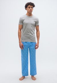 Νεαρός άνδρας που στέκεται ξυπόλητος, φορώντας γκρι μπλουζάκι και μπλε παντελόνια πιτζάμας με άσπρα σχέδια αλόγου, σε απλό λευκό φόντο.