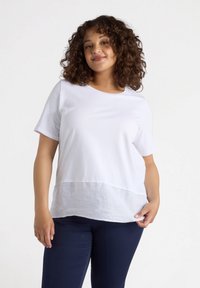 Biała bluzka z krótkim rękawem, o okrągłym dekolcie, z lekkim, teksturowanym dolnym panelem i gładką górną częścią. Połączona z ciemnymi spodniami.