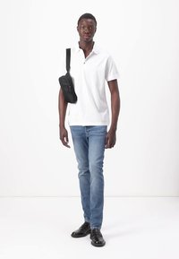 Witte polo, blauwe jeans, zwarte schoudertas. Model staat op een witte achtergrond. Schoenen zijn zwart met een gladde afwerking.