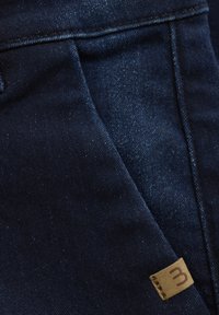 Mörk denimtyg med en subtil lyster, med en liten tanfärgad läderetikett sydd på sidan och en strömlinjeformad, modern passform.