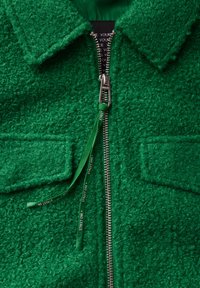 Grön fleecejacka med full dragkedja, krage, två framtida fickor och texturerad finish. Inkluderar en märkeslogotyp på dragkedjan.