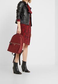 Punainen reppu, jossa on teksturoitu kuvio ja kultaiset vetoketjuyksityiskohdat. Yhdistetty punaisen kuviollisen mekon ja mustien niittikorkokenkien kanssa.