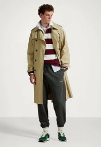 Άνδρας που φοράει μπεζ παλτό πάνω από μπλούζα με μπορντό και λευκές ρίγες, σκούρες γκρι φόρμες, πράσινα αθλητικά παπούτσια, στέκεται με το ένα χέρι στην τσέπη.