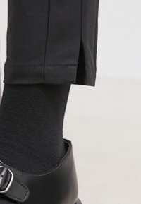 Gros plan sur le bas d'un pantalon noir avec une fente latérale, porté sur une chaussette noire et une chaussure en cuir noir brillant avec boucle.