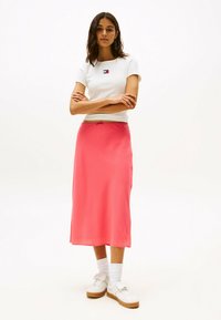 Hvid tætsiddende t-shirt med lille logo, parret med en mellemlang, lys pink satin nederdel. Hvide sneakers med en lys tan sål.