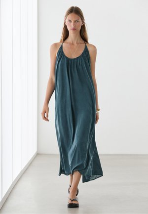 Femme portant une robe maxi ample bleu sarcelle à col halter, marchant à l’intérieur sur un sol gris clair contre un mur blanc.