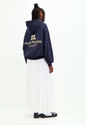 Frau mit geflochtenem Haar, die einen navyblauen Hoodie mit dem Schriftzug „Pacific Republic“, einen weißen Stufenrock und blaue Sneaker trägt, steht vor einem weißen Hintergrund.