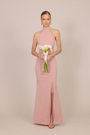 Femeie într-o rochie fără mâneci, roz pal, cu guler înalt și șliț, ținând crini albi legați cu panglică, purtând bijuterii aurii și sandale.