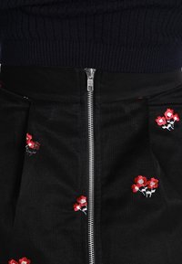 Černá texturovaná sukně se stříbrným předním zipem a malými vyšitými červenými a bílými květinovými menšími shluky poblíž zipu pod tmavým žebrovaným topem.