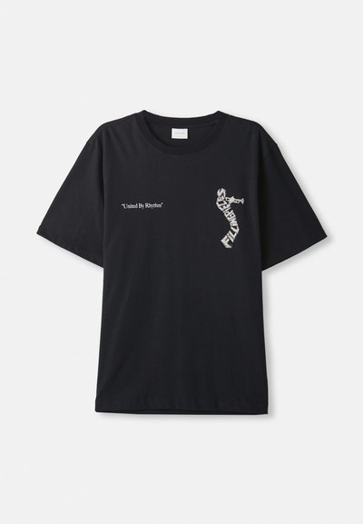 Svart bomullströja med oversized passform. Har vit text "United By Rhythm" och grafisk design på framsidan. Klassisk rundhalsad stil.