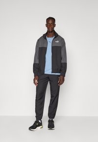 Schwarz-graue Zip-Jacke mit elastischen Bündchen, getragen über einem hellblauen Hemd, kombiniert mit schwarzen Hosen und schwarzen Laufschuhen.