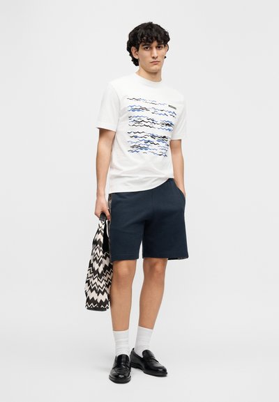 Λευκό T-shirt με γραφικό κύματος σε μπλε και μαύρο, συνδυασμένο με ναυτικό σορτς. Το μοντέλο κρατάει μια τσάντα με ασπρόμαυρο ζιγκ-ζαγκ σχέδιο.