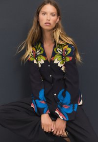 Chemise à manches longues en tissu foncé présentant des motifs floraux audacieux en vert, bleu et rouge, avec une patte de boutonnage sur le devant et une coupe décontractée.