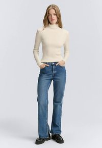 Cremefarbener, gerippter Rollkragenpullover kombiniert mit blauen ausgestellten Jeans, die eine hohe Taille, dezente Nähte und schwarze Plateauschuhe aufweisen.