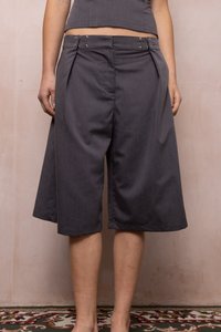 Person iført mørkegrå vidde knælange bukser med metalknapper i taljen, stående på mønstret tæppe mod en lys væg.