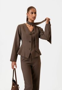 Hnedý prispôsobený oblek s priliehav�ým sakom s V-výstrihom, tromi gombíkmi a viazanou šatkou ako akcentom. Nohavice sú rovné, v rovnakom farebnom odtieni.
