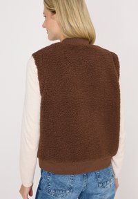 Brown fleece vest featuring a textured, fluffy exterior, ribbed collar, and hem. Worn over a light long-sleeve top and blue jeans.