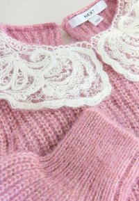 Różowy sweter z dzianiny o teksturowanym tułowiu i białym kołnierzyku z koronką, ozdobionym finezyjnymi wzorami. Metka widoczna przy szyi.