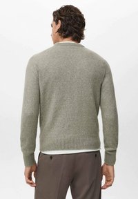 Hombre con cabello ondulado que lleva un suéter de manga larga gris claro y pantalones marrones, de pie de espaldas frente a un fondo liso.