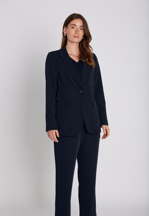 Jeune femme aux longs cheveux bruns portant un costume bleu marine foncé avec blazer et pantalon assorti, debout devant un fond gris clair.
