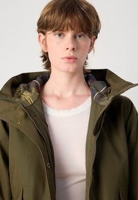 Olijfgroene jas met een ritssluiting aan de voorkant, voorzien van een patroon aan de binnenvoering die zichtbaar is bij de capuchon. Het model draagt eronder een witte geribde shirt.