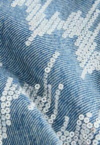 Denimstof med en lys blå vask, der har indviklede mønstre af hvide perler arrangeret i bølgelignende designs hen over overfladen.