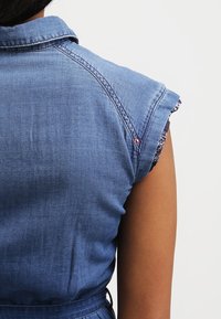 Koszula dżinsowa z krótkimi, podwiniętymi rękawami, w kolorze jasnoniebieskim, widoczne detale szycia, podkreślona wzorzystym materiałem wystającym z mankietów.