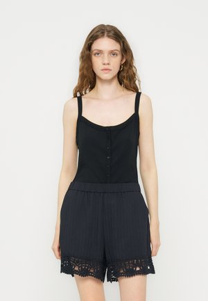 Nuori nainen, jolla on aaltoilevat hiukset, pukeutunut hihattomaan mustaan napitettuun toppiin ja mustiin pitsireunaisiin shortseihin, seisoo vaalean yksivärisen taustan edessä.