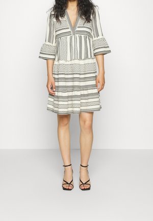 Nainen pukeutuneena polvipituiseen valkoiseen mekkoon, jossa on mustia geometrisia kuvioita ja levenevät kolmen neljänneksen hihat, yhdistettynä mustiin remmikkäisiin korkokenkiin.