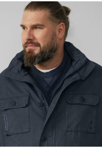 Donker marineblauwe jas met een volledige rits, twee borstzakken met kleppen en een gestructureerde afwerking. Casual, met een capuchon en knoopdetails.