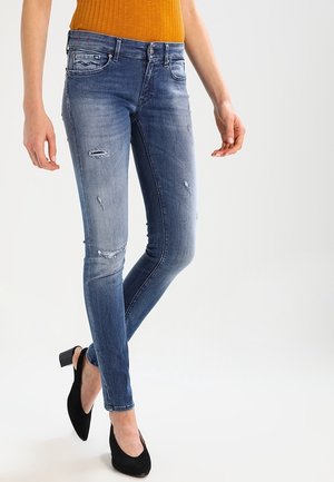 Personne portant un jean skinny bleu déchiré, un haut côtelé orange et des chaussures slingback noires à talon bloc, debout, jambes croisées.